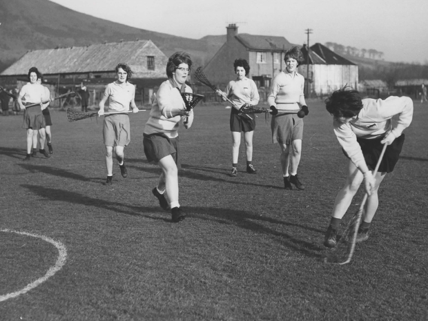 Historic black-and-white photograph of a group of women playing lacrosse on an outdoor pitch. One player is passing the ball to another, while their teammates are running behind them.
