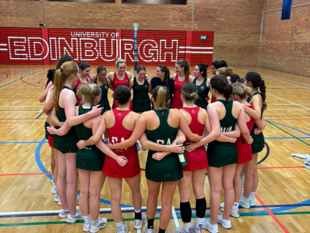 A group of netball players stand on the middle of a netball court in a huddle, dressed in green and red netball dresses.