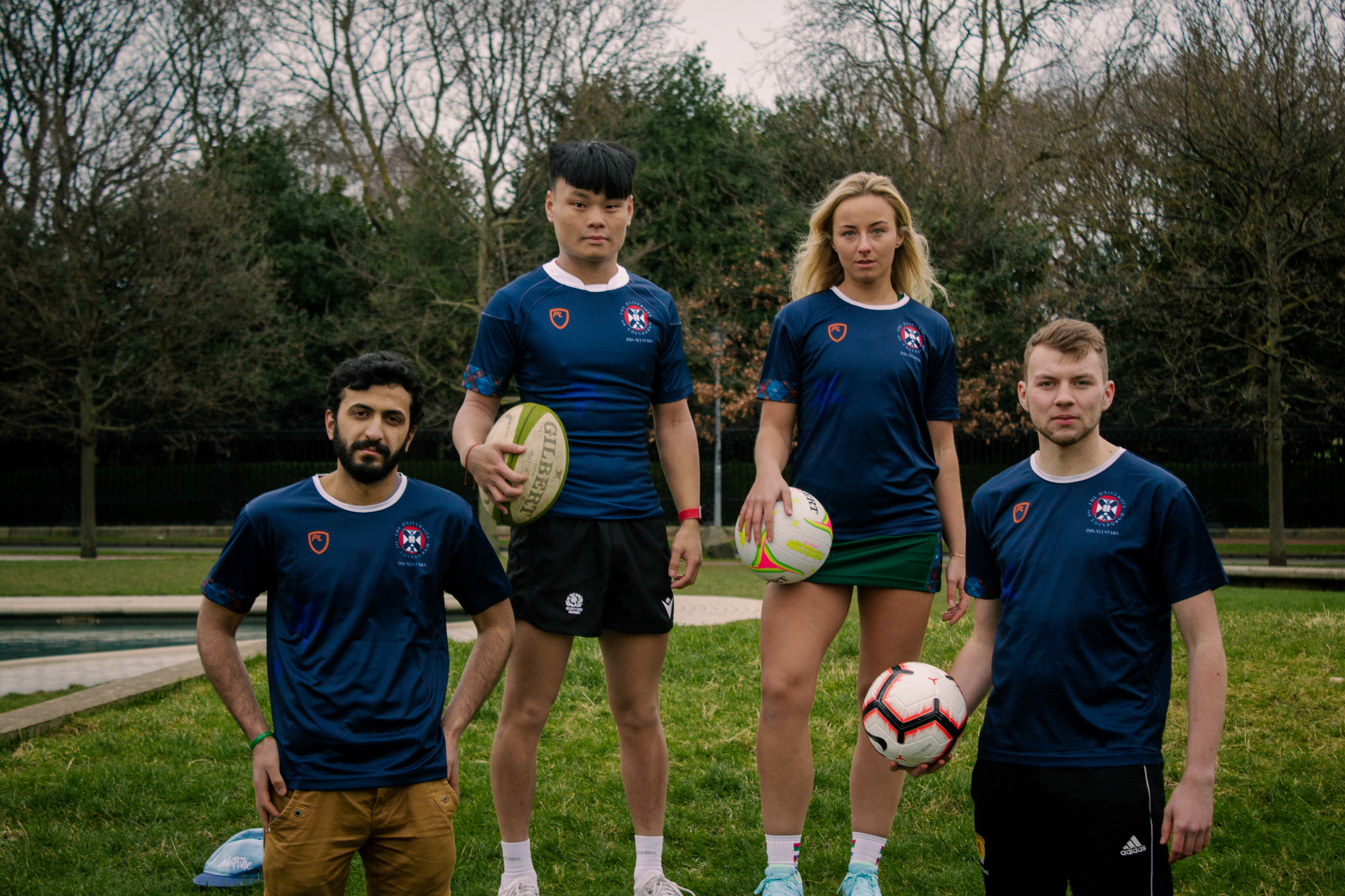 Four students posing wearing dark blue t-shirts, holding a rugby ball, a netball, and a football
