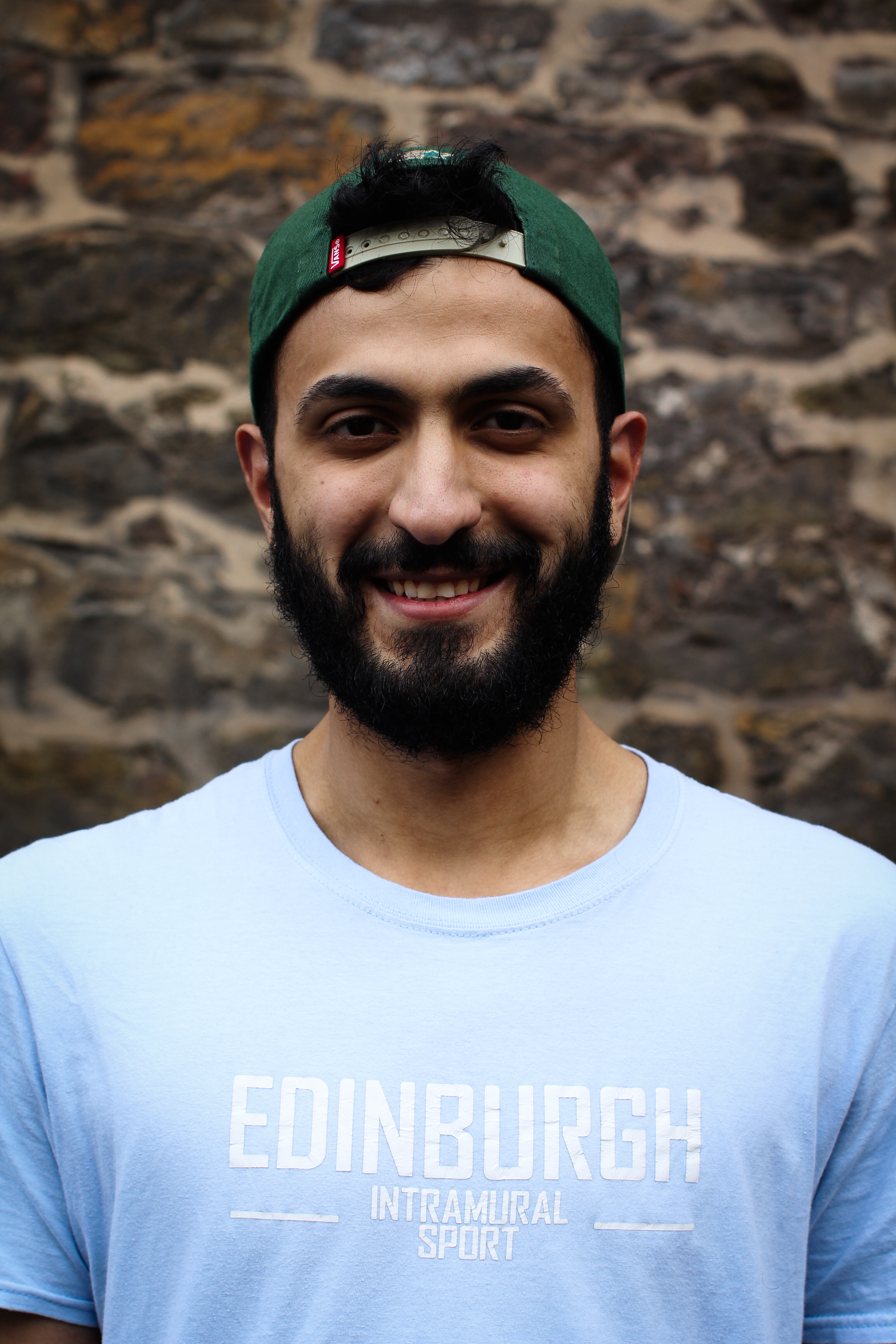 Abd wearing a light blue t-shirt with Edinburgh intramural sport written on the front in white writing, and wearing a green cap backwards on his head.