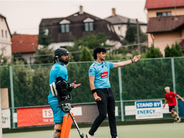 An outdoor field hockey match on an artificial turf pitch. A goalkeeper in protective gear stands near the goal while an official in a light blue shirt signals with an outstretched arm.