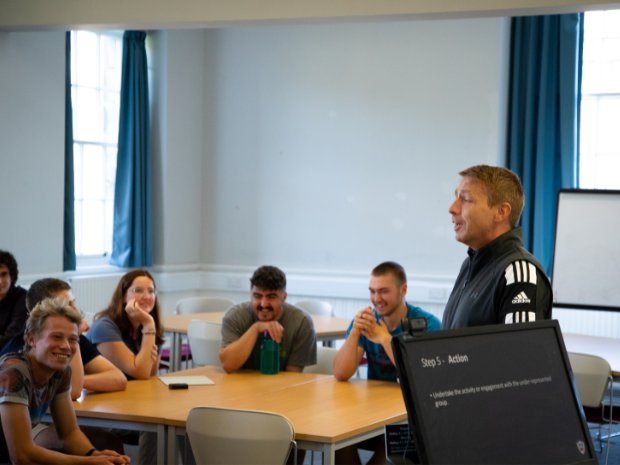 A facilitator stands at the front of a classroom-style training room speaking to a group of participants seated around tables.