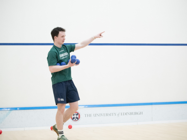 A participant holds blue boccia balls while pointing across the court during a coached session. Red and blue balls are visible on the floor of the indoor sports hall.