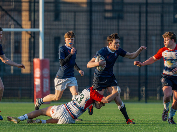 Intramural Sport participants competing in a rugby match on an outdoor sports pitch, with one player carrying the ball while being tackled by an opposing player.