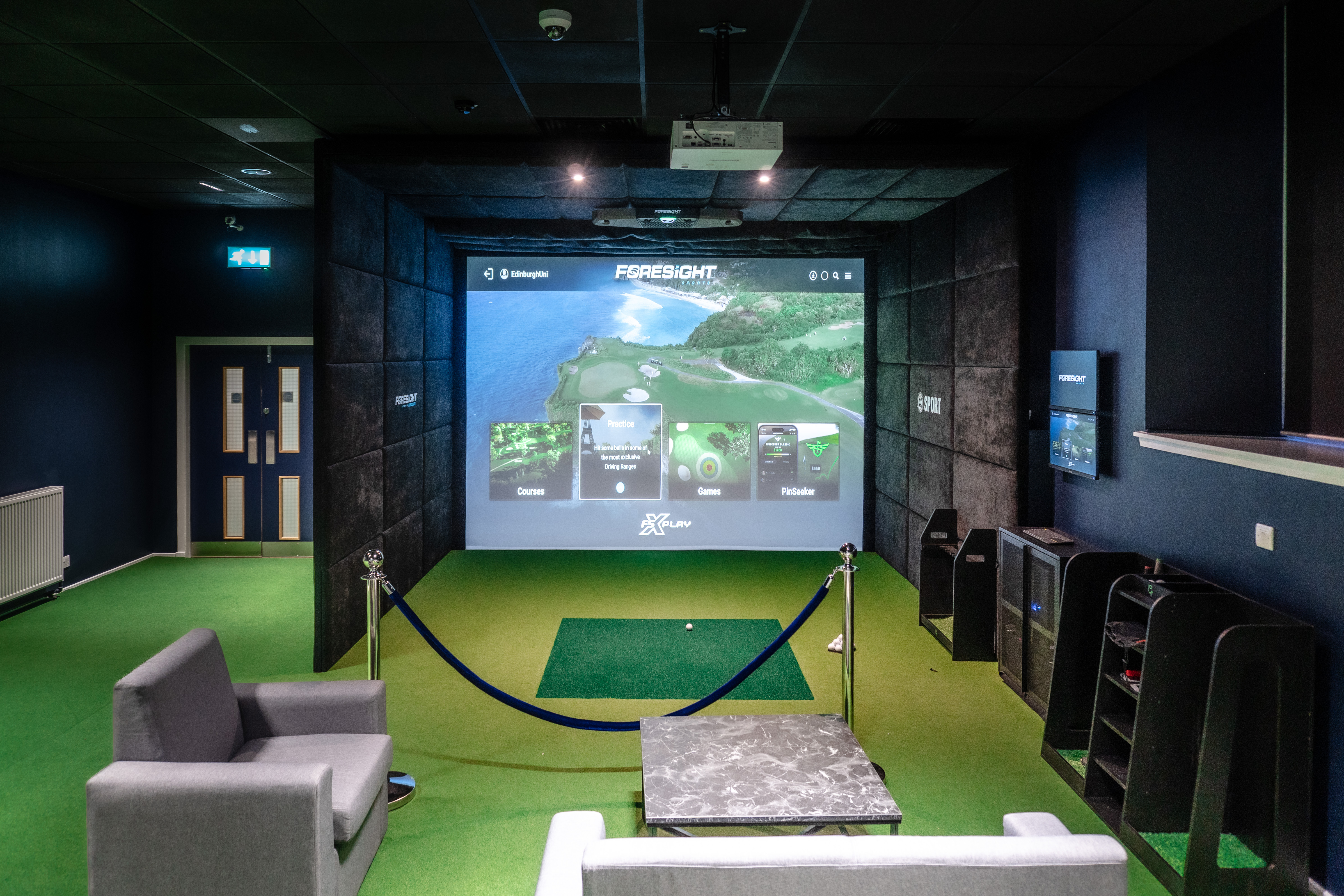 Golf simulator. Large screen showing optional courses to play with a mat in front of it and a golf ball. Behind the playing area is a small seating area which is cordoned off. 