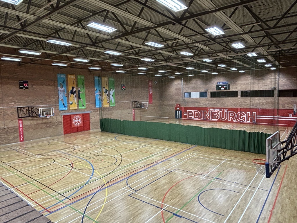 Indoor Sports hall, six badminton court size, lined for various sports, with drop down netting width ways across the hall at halfway. Walls of hall are brick, with Edinburgh Sport branding spread throughout the venue. 