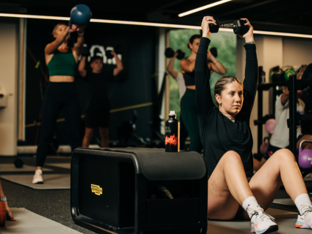 Busy gym space with people using kettle bells and dumbells, lighting focussed on woman doing weighted sit ups next to a technogym bench